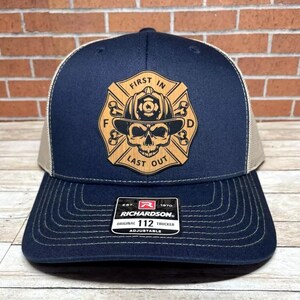 May include: A navy blue and tan trucker hat with a wooden patch featuring a skull with crossed bones and the text "First In Last Out". The patch is in the shape of a fire department badge. The hat has a Richardson 112 adjustable strap.