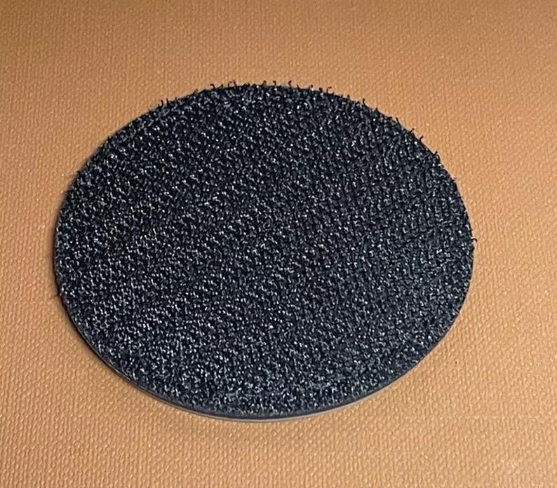 May include: A round, black, adhesive-backed hook and loop fastener. The fastener is made of a soft, fuzzy material.