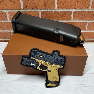 May include: Black and tan pistol patch with a black magazine on a brown box. The patch has a detailed design with a red dot sight and a light attachment. The magazine is black and has a brass-colored tip.