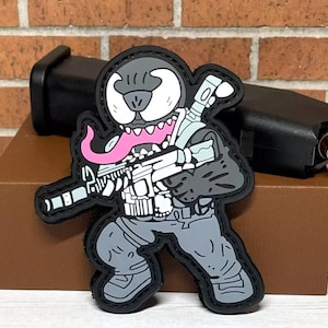 May include: A black and gray patch with a cartoon character resembling Venom holding a rifle. The character has a pink tongue and a black and white face.