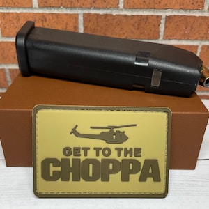 May include: A tan fabric patch with a black silhouette of a helicopter and the text "GET TO THE CHOPPA" in brown letters. The patch is sitting on top of a brown box.