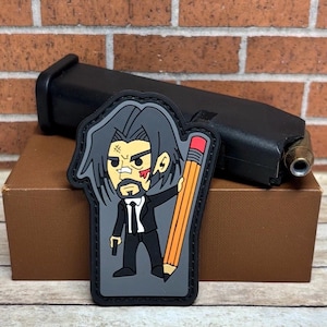 May include: A black and gray patch featuring a cartoon character in a suit holding a large pencil. The patch is placed in front of a black pistol magazine and a brown box, with a brick wall in the background.