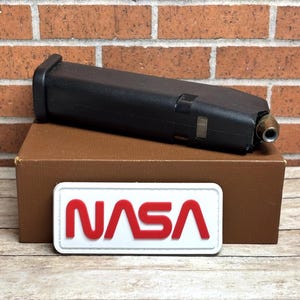 May include: A black magazine with a brass-colored tip rests on a brown box. A white patch with the red letters "NASA" is in the foreground. The background is a brick wall.