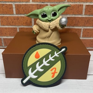 May include: A green and white Mandalorian symbol patch with a red drop of blood and the number 7 in orange. The patch is on a brown surface.