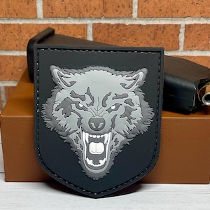 May include: Black patch with a gray wolf head with a snarling expression. The wolf's eyes are white and the patch has a stitched border.