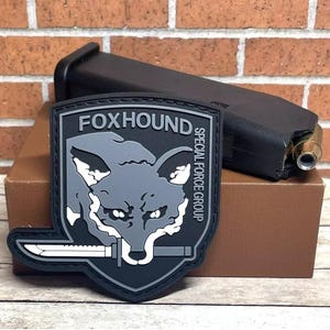 May include: A black and gray patch with a fox head and the text "FOXHOUND SPECIAL FORCE GROUP".