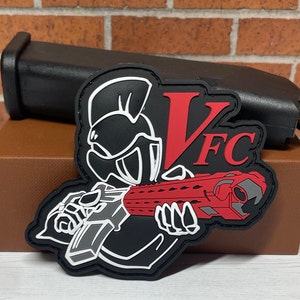 May include: Black and white patch with a red and gray gun, a white helmet, and the text "VFC" in red.