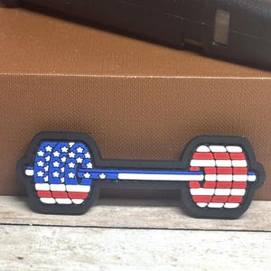 May include: A black dumbbell-shaped patch with a patriotic design. The ends feature the American flag, with red and white stripes and white stars on a blue background. A blue and white stripe runs through the center.