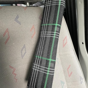 May include: A rectangular seat belt cover featuring a black, white, and green plaid pattern. The fabric accessory is wrapped around a black seat belt strap, positioned against a light grey car seat with a subtle geometric pattern in red and blue.