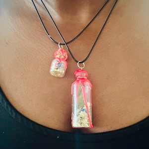 May include: Two glass bottle pendant necklaces. The bottles are filled with small, colourful objects and sealed with red wax. The necklaces have black cords and silver-coloured clasps. The pendants are different sizes.