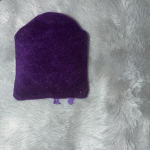 Puede incluir: Un expositor de joyas de terciopelo morado oscuro con una parte superior redondeada y una base rectangular. El expositor est&aacute; sobre un fondo esponjoso de color gris claro.