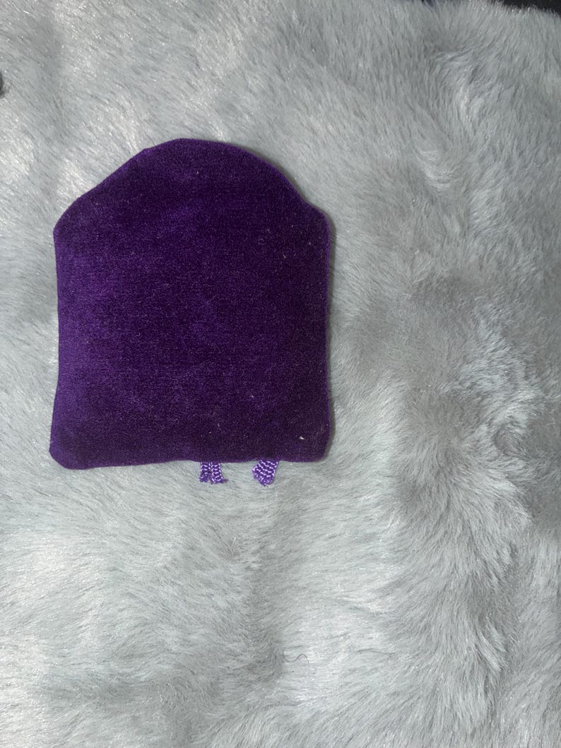 Puede incluir: Un expositor de joyas de terciopelo morado oscuro con una parte superior redondeada y una base rectangular. El expositor est&aacute; sobre un fondo esponjoso de color gris claro.