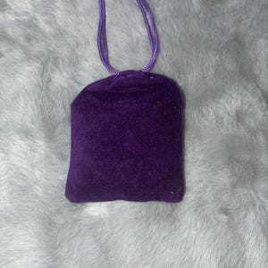 Puede incluir: Una peque&ntilde;a bolsa cuadrada de terciopelo morado oscuro con cord&oacute;n. La bolsa tiene un cierre de cord&oacute;n morado a juego. La bolsa est&aacute; sobre una superficie esponjosa de color gris claro.