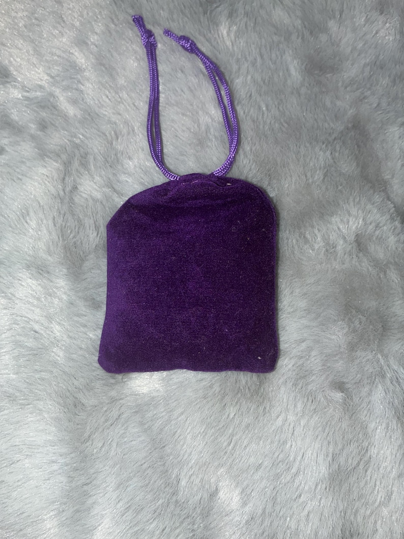 Puede incluir: Una peque&ntilde;a bolsa cuadrada de terciopelo morado oscuro con cord&oacute;n. La bolsa tiene un cierre de cord&oacute;n morado a juego. La bolsa est&aacute; sobre una superficie esponjosa de color gris claro.