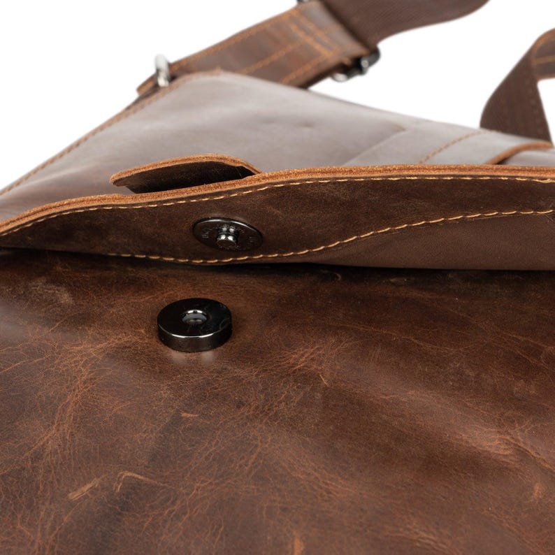 May include: Close-up of a brown leather messenger bag with a magnetic closure. The bag has a flap with a stitched edge and a metal snap closure.