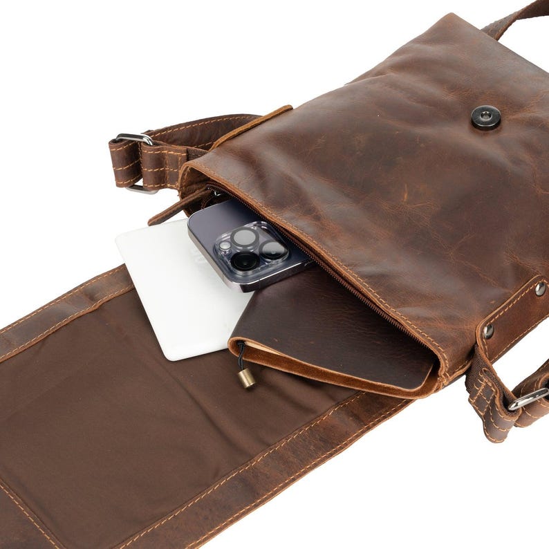 May include: A brown leather messenger bag with a flap closure and a zippered pocket. The bag is open and has a phone and a notebook inside.