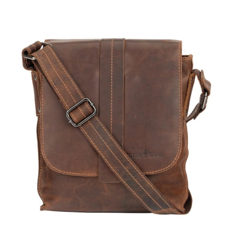 May include: A brown leather messenger bag with a long adjustable strap and a buckle closure. The bag has a flap with a stitched design and a small pocket on the front. The bag has a brand label that says "Leather & Wood".