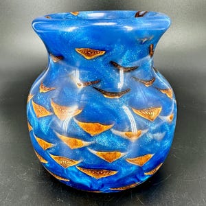 Peut inclure: Un vase bleu avec une finition chatoyante et scintillante. Le vase est orné de nombreux accents bruns en forme de feuilles. Le vase a une base large et un bord légèrement évasé.
