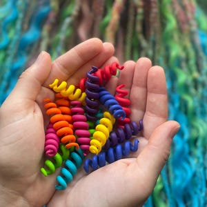 Könnte beinhalten: Ein Paar Hände hält eine Sammlung farbenfroher, spiralförmiger Haaraccessoires. Die Accessoires sind in Blau-, Grün-, Gelb-, Orange-, Rosa- und Lilatönen gehalten. Der Hintergrund zeigt lange, lockige Haare in ähnlichen leuchtenden Farben.
