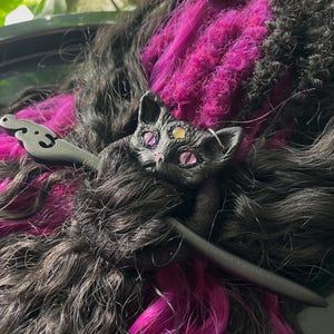 May include: A black cat hair accessory with pink eyes and a third eye, resting on black and pink hair extensions. The accessory is a decorative hair piece, possibly a barrette or hair clip, with a stylized cat design.