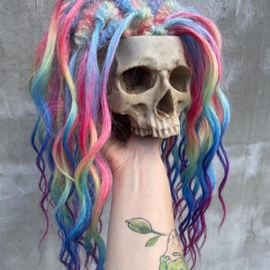 May include: A beige human skull planter adorned with rainbow-coloured dreadlocks. The hair features shades of pink, blue, yellow, and green. The dreadlocks cascade down, creating a unique and artistic display. A striking and unusual decorative item.