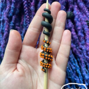 May include: A wooden skewer decorated with black and orange polymer clay tentacles. The top portion is black and coiled, while the lower section features orange suction cups. The text "Set of 2 (#2)" is in a white circle.