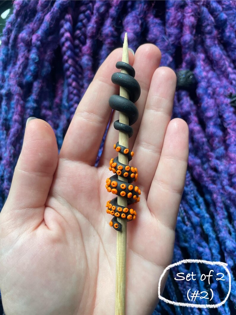 May include: A wooden skewer decorated with black and orange polymer clay tentacles. The top portion is black and coiled, while the lower section features orange suction cups. The text "Set of 2 (#2)" is in a white circle.