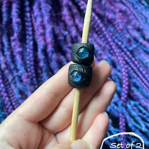 May include: Two black square beads with blue iridescent centers are stacked on a wooden skewer. The beads have a textured surface. The background is a blurred pattern of purple and blue.