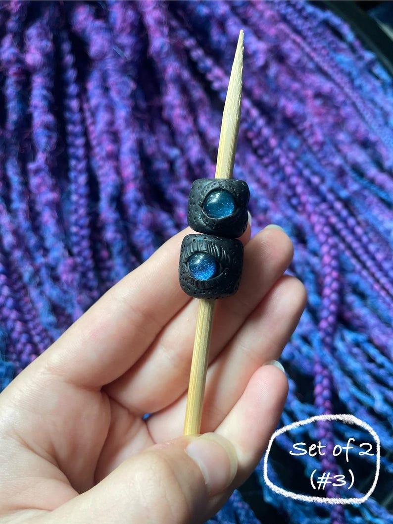 May include: Two black square beads with blue iridescent centers are stacked on a wooden skewer. The beads have a textured surface. The background is a blurred pattern of purple and blue.