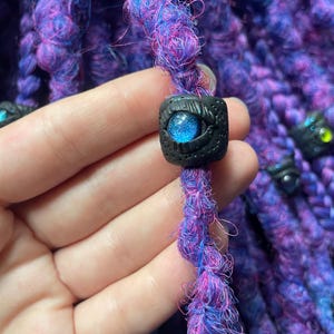 May include: Close-up of purple and blue dreadlocks with a black bead featuring a blue eye design. The dreadlocks are a mix of purple and blue fibers, with a textured appearance. The bead is square-shaped with a detailed eye design.