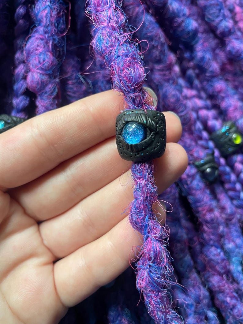 May include: Close-up of purple and blue dreadlocks with a black bead featuring a blue eye design. The dreadlocks are a mix of purple and blue fibers, with a textured appearance. The bead is square-shaped with a detailed eye design.