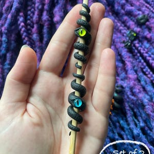 May include: A wooden hair stick with a black, coiled snake design. The snake has textured scales and two glowing eyes, one green and one blue. The text "Set of 2 (#1)" is in a white circle.