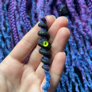 May include: A blue and purple dreadlock extension with a black, coiled snake charm. The snake charm has a bright green eye. The background features more blue and purple dreadlocks.
