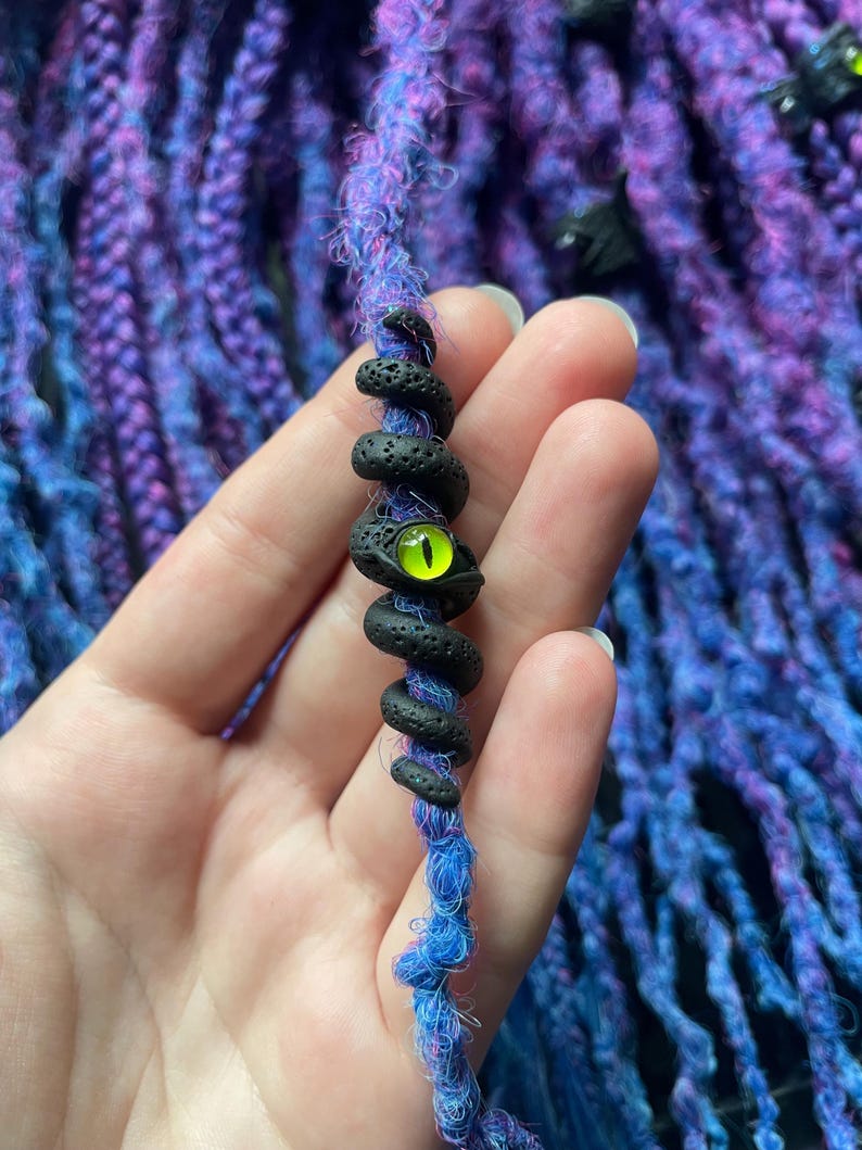 May include: A blue and purple dreadlock extension with a black, coiled snake charm. The snake charm has a bright green eye. The background features more blue and purple dreadlocks.