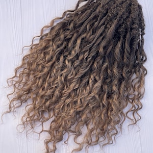 May include: A collection of brown and blonde curly hair extensions. The hair features long, loose curls with a gradient effect, shifting from dark brown at the roots to a lighter blonde at the tips. The hair is placed on a white wooden surface.