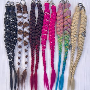 Könnte beinhalten: Acht verschiedene geflochtene Haarverlängerungen in verschiedenen Farben und Verzierungen, darunter Weiß, Braun, Pink, Blau und Blond. Jede Verlängerung hat einen Metallring oben zum Anbringen an einem Schlüsselanhänger oder einem anderen Objekt.