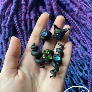 May include: A set of five black polymer clay rings and sculptures, each featuring a unique eye design with blue and green irises. The pieces are held in a hand against a backdrop of purple and blue fabric.