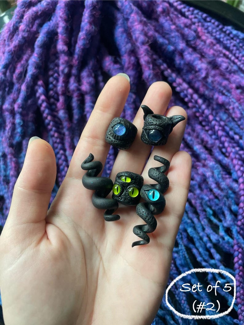 May include: A set of five black polymer clay rings and sculptures, each featuring a unique eye design with blue and green irises. The pieces are held in a hand against a backdrop of purple and blue fabric.
