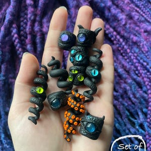 May include: A set of ten black polymer clay charms with various eye designs. Some charms have horns or tentacles. The eyes are blue, green, and purple. The charms are held in a hand.