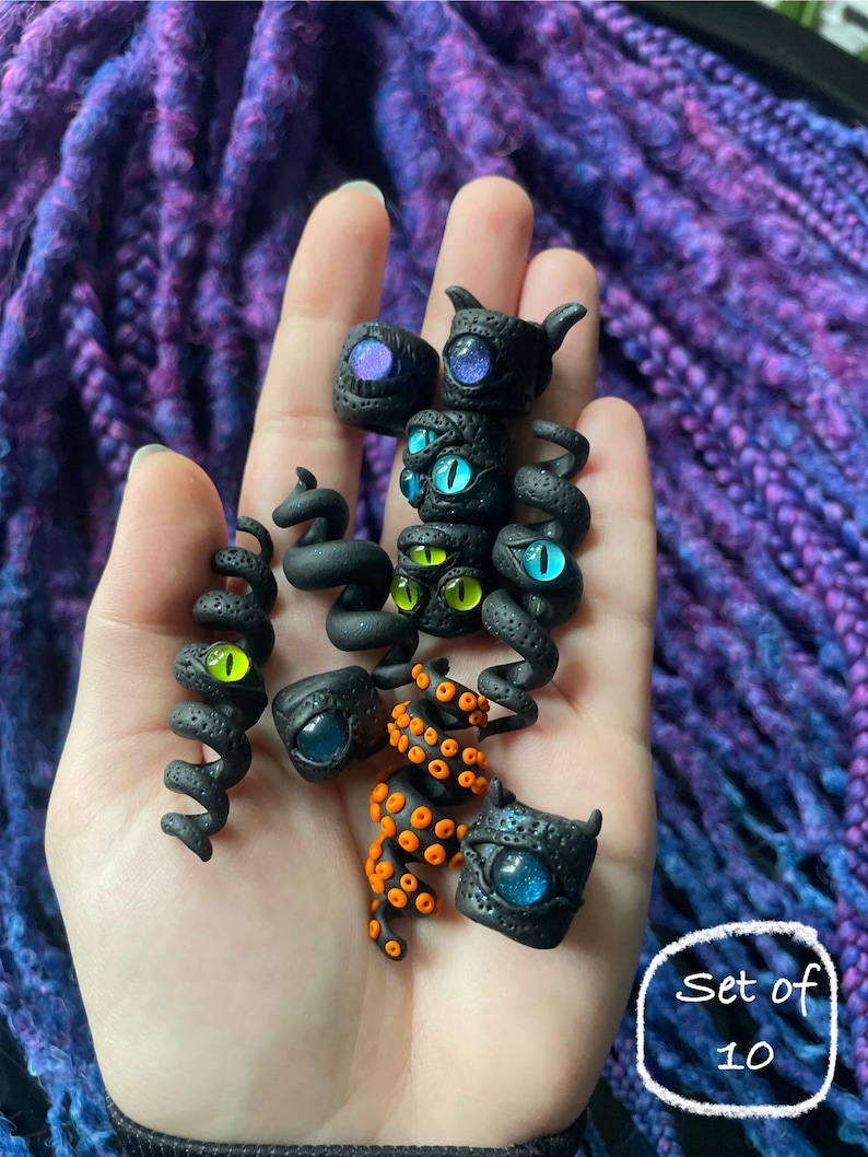 May include: A set of ten black polymer clay charms with various eye designs. Some charms have horns or tentacles. The eyes are blue, green, and purple. The charms are held in a hand.