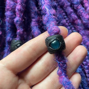 May include: Close-up of a vibrant purple and blue dreadlock with a black bead featuring a blue eye design. The dreadlock is made of textured, colorful yarn. The bead is square-shaped and has a detailed eye. The background is a similar dreadlock.