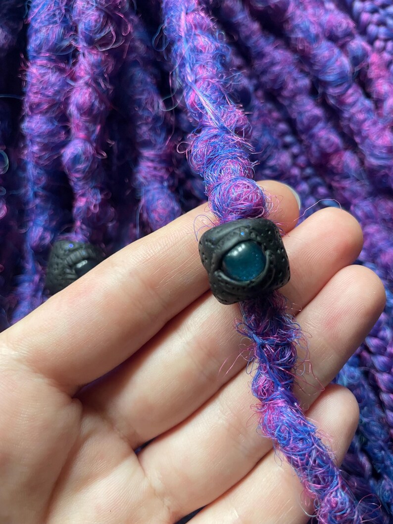 May include: Close-up of a vibrant purple and blue dreadlock with a black bead featuring a blue eye design. The dreadlock is made of textured, colorful yarn. The bead is square-shaped and has a detailed eye. The background is a similar dreadlock.
