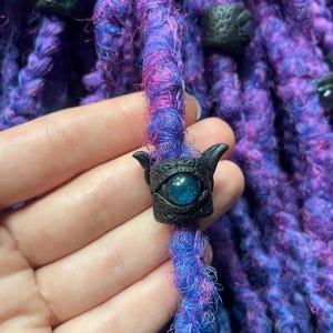 May include: Close-up of purple and blue dreadlocks with black decorative beads. The beads have an eye design with a blue glittery center. The dreadlocks are a mix of purple, blue, and pink fibers.