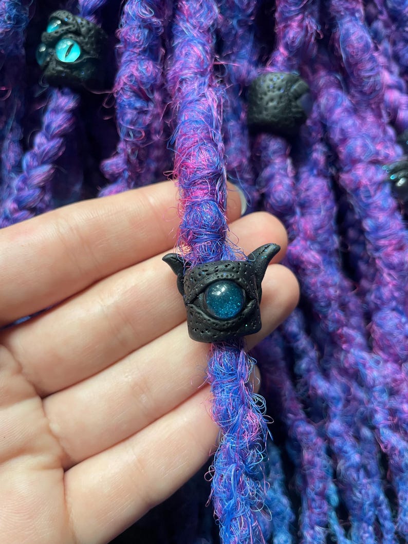 May include: Close-up of purple and blue dreadlocks with black decorative beads. The beads have an eye design with a blue glittery center. The dreadlocks are a mix of purple, blue, and pink fibers.