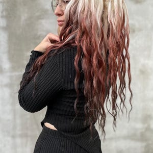 Pode incluir: Uma pessoa com cabelo longo e ondulado em tons de branco, vermelho e castanho. A pessoa está vestindo um top e uma saia de malha canelada preta. Eles também estão usando óculos redondos com armação de arame.