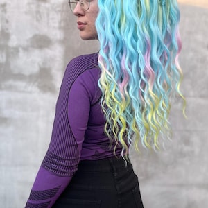 May include: A person wearing a purple long-sleeve shirt with black jeans. The shirt has black patterned designs on the sleeves. The person has blue, yellow, and pink wavy hair.