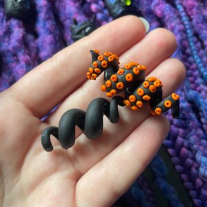 May include: A black and orange octopus-shaped hair accessory. The accessory features a coiled body and tentacles with orange suckers. The background is a mix of purple and blue, suggesting a handmade craft item.