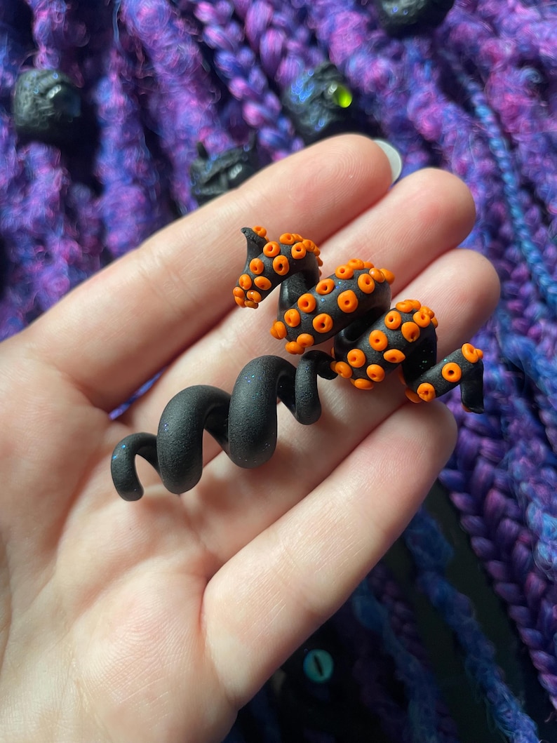 May include: A black and orange octopus-shaped hair accessory. The accessory features a coiled body and tentacles with orange suckers. The background is a mix of purple and blue, suggesting a handmade craft item.