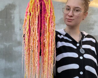 May include: A set of long, colorful synthetic dreadlocks in shades of pink, yellow, and white. The dreadlocks are held up by a person wearing a black shirt with a white skeleton design.