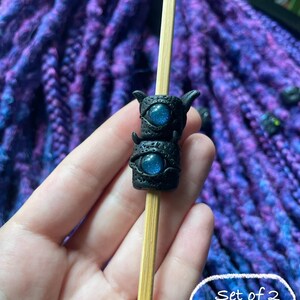 May include: A pair of black, horned dreadlock beads with blue, glittery eye details are displayed on a wooden stick. The beads are stacked, and the background features purple and blue dreadlocks. The image includes the text "Set of 2 (#4)".